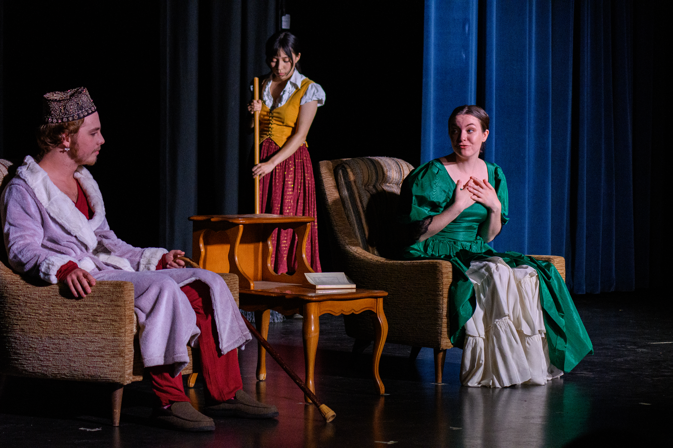 Three actors on stage. Two are sitting in armchairs. The third is sweeping.